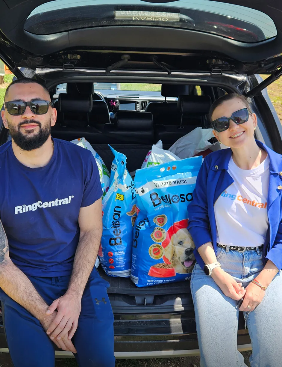Двама служители на RingCentral, седнали на отворения багажник на автомобил, пълен с чували с храна за кучета.