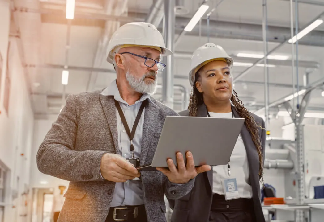 Two workers wearing hard hats engaged in a discussion while looking at a laptop